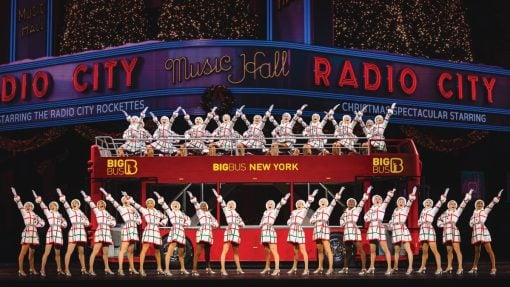 The rockettes stand in front of a big red bus outside of radio city