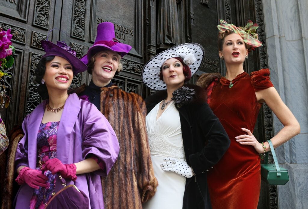 women in colorful dresses, big hats, and accesorries pose in front of a door