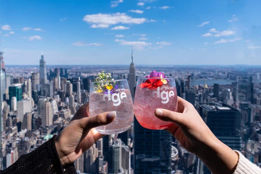 two hands toasting cocktail in front of nyc skyline at the edge in new york