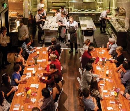 Two long tables are full of patrons eating as they look into the open kitchen at Lazy Bear