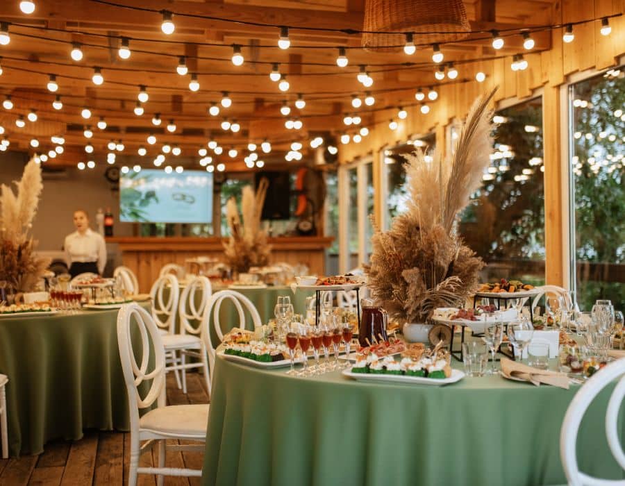 Large group holiday party ideas with a beautifully decorated venue featuring round tables set with drinks and appetizers under ambient string lights. This photo depicts a spacious, inviting setting for large gatherings, ideal for companies planning a holiday party that accommodates high-capacity events in a festive and elegant atmosphere.