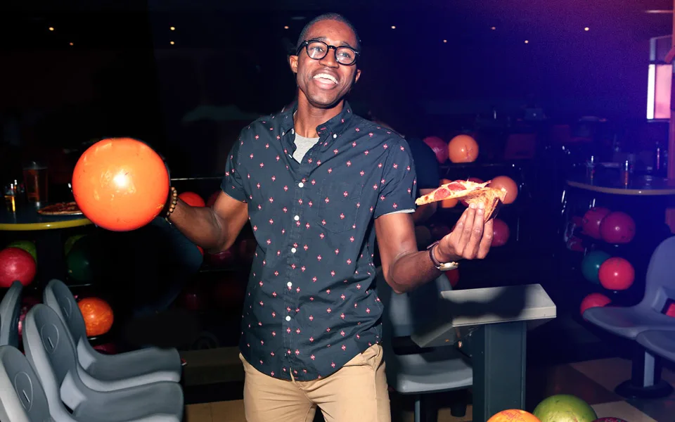 A man with pizza in one hand and a bowling ball in the other poses