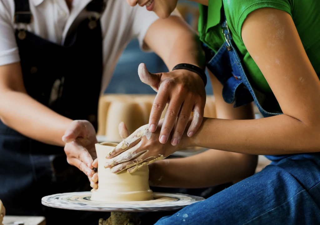 An instructors hands guide a students in making a vessel on a pottery wheel