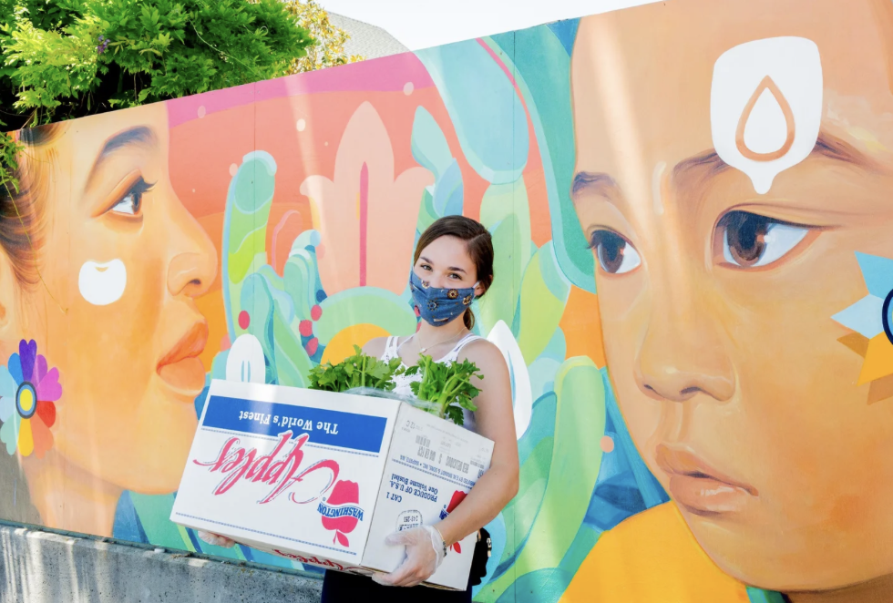 A woman wearing PPE holds a box of fresh produce