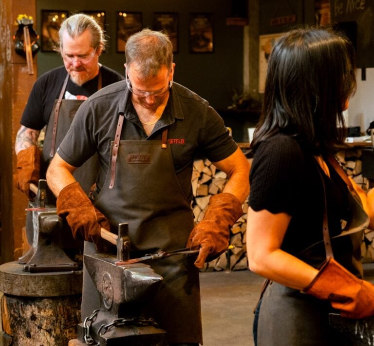 A man with goggles looks intently at the metal he's welding in the forge