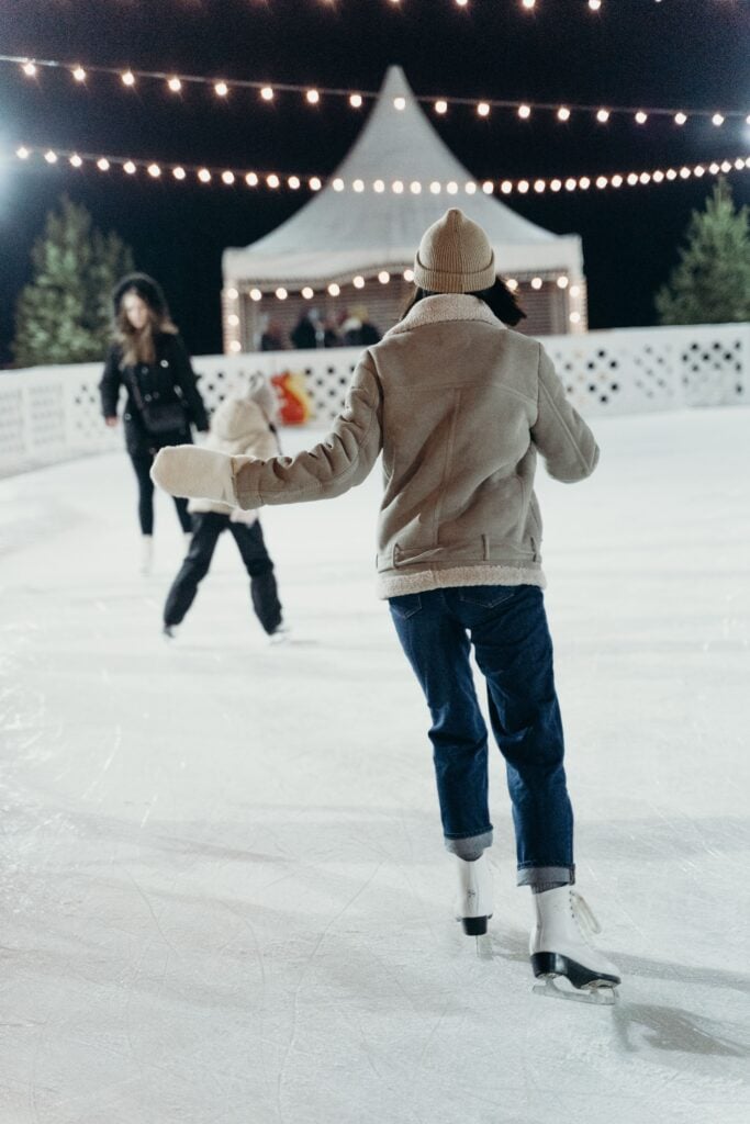 A woman is ice skating down the center of the link as string lights glow overhead