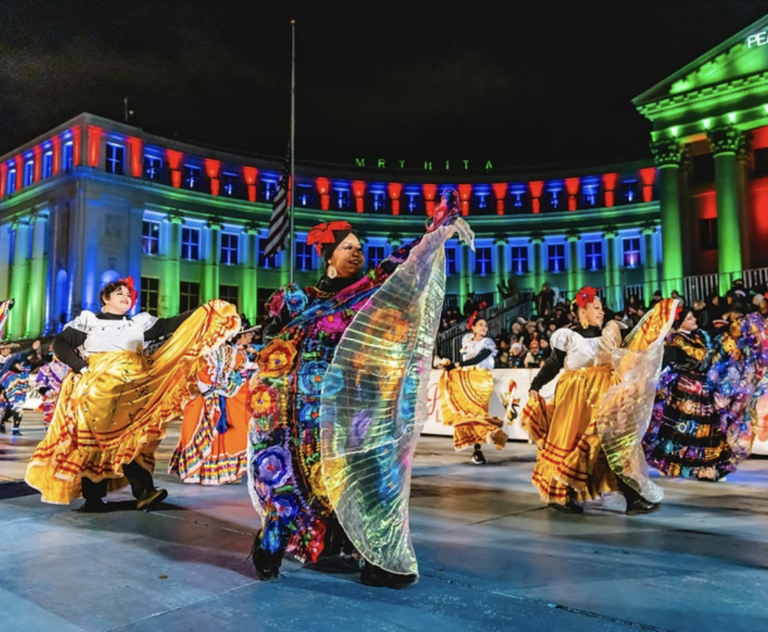 Women in beautoul costumes dance in front o lighted buildings
